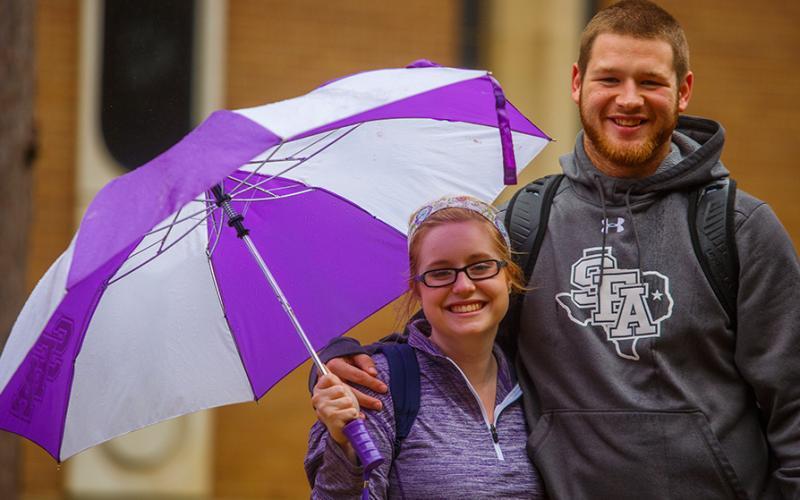 Two graduate students wearing SFA attire stand together with a purple and white umbrella