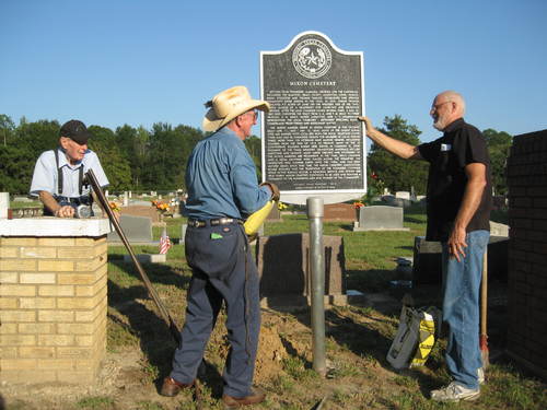 Cherokee County Historical Commission members, Shelley Cleaver and John Thomason place the marker in the ground while cemetery president, Elray Partin, looks on.