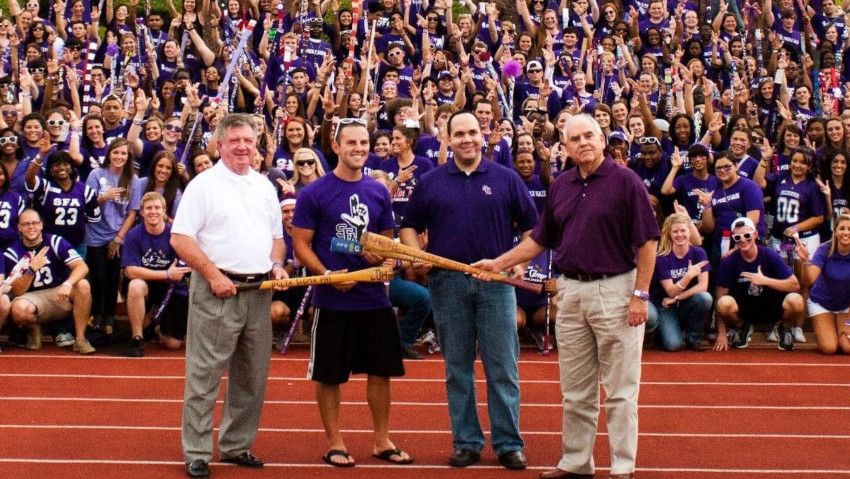Sawyers and SFA students, holding axe handles, stand in front of a large crowd. Members of the assembly are showing the axe 'em hand sign.