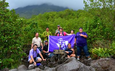 Students, faculty members, and a local guide pose for a photo in front of Arenal Volcano, obscured by cloud cover.