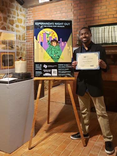 Ajani Brown holds the Mitchell A. Wilder Silver Publication Design Award while standing next to his marketing poster