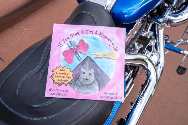 The cover of Katherine Noble’s book, "If You Give a Girl a Motorcycle," depicts a cat, a highway, and handlebars. The book rests on a motorcycle seat.