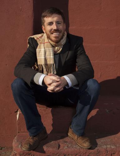 Comedian Joe Zimmerman crouches in front of a rust-red brick wall 