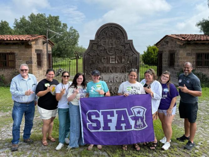 Graduate students, representatives from Hacienda Santa Clara, and SFA faculty members hold an SFA flag in front of a landmark in San Miguel de Allende