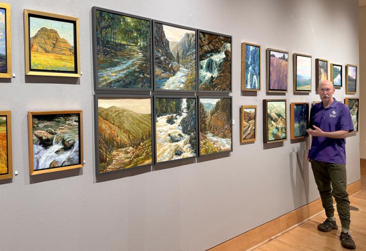 Faculty artist Peter Andrew stands in front of a gallery wall featuring his paintings