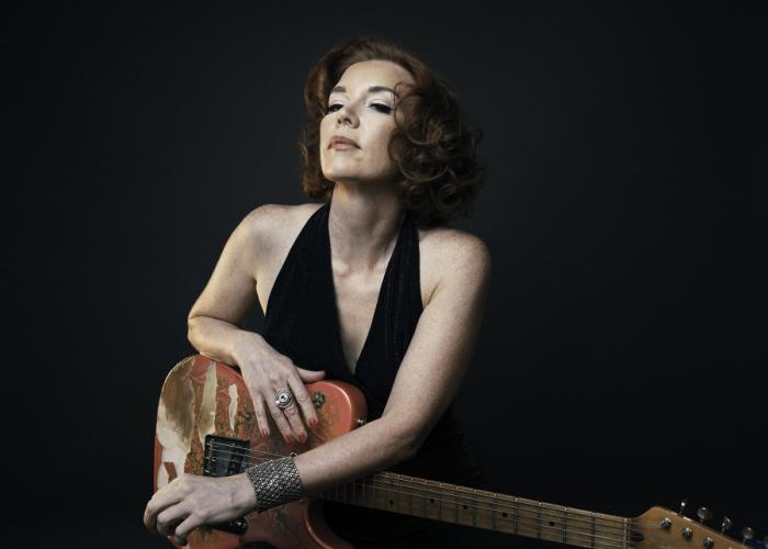 Sue Foley, set against a dark backdrop, holds an electric guitar 