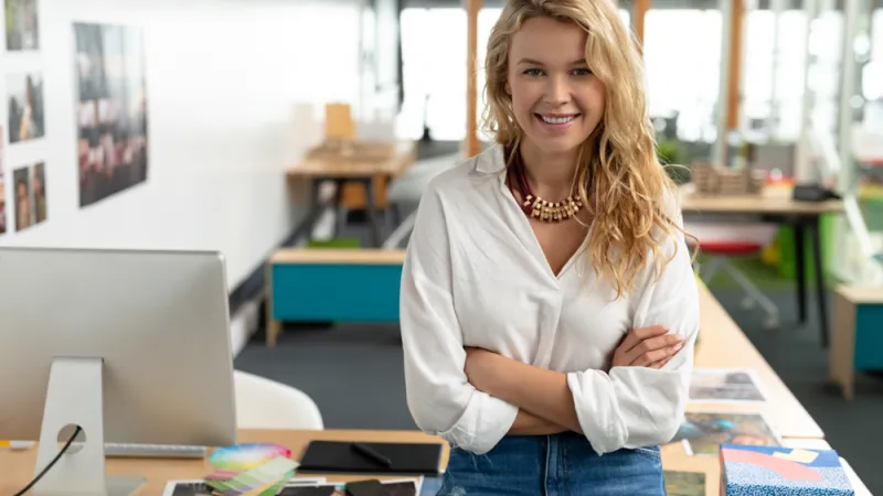 Portrait of Caucasian female graphic designer with arms crossed sitting at desk in a modern office. This is a casual creative start-up business office for a diverse team
