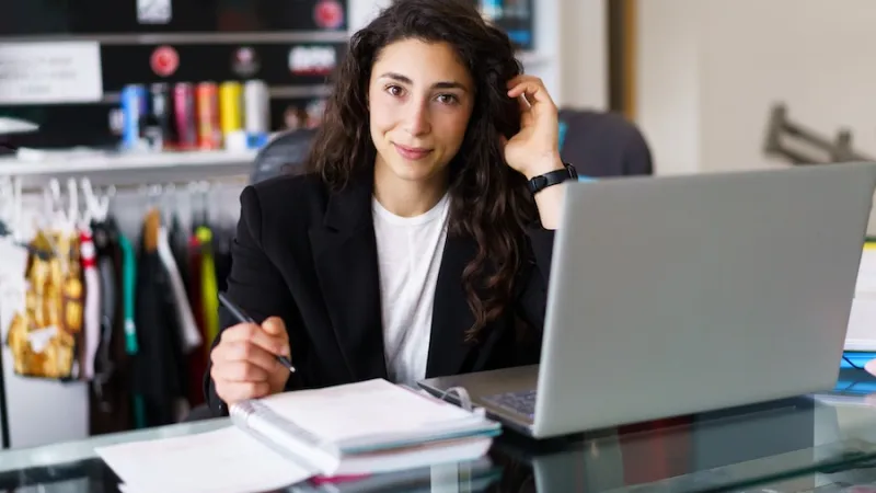 Positive brunette looking at camera and adjusting long wavy hair while sitting at desk with netbook and making notes during psychology session