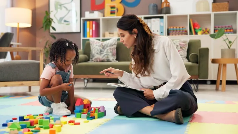 Therapist, woman and girl with clipboard, blocks and talk for evaluation, assessment or exam on floor. Person, psychologist and child with toys, advice and checklist for notes with point at clinic