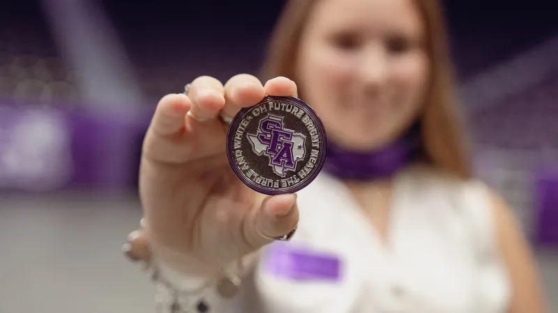 Hailey Crews displays the silver challenge coin she received. The coin displays the text, "Oh future bright 'neath the purple and white."
