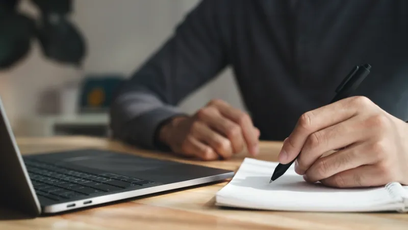 Closeup of man writing with a pen on paper in front of a laptop