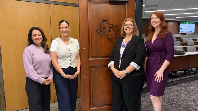 Four SFA alumnae are pictured standing in front of a wooden door stamped with the SFA logo