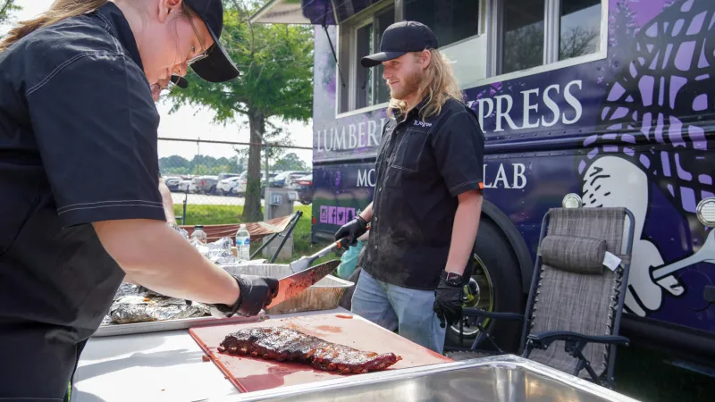 Axes and Embers team members prepare barbecue on a table next to the Lumberjack Express mobile food lab