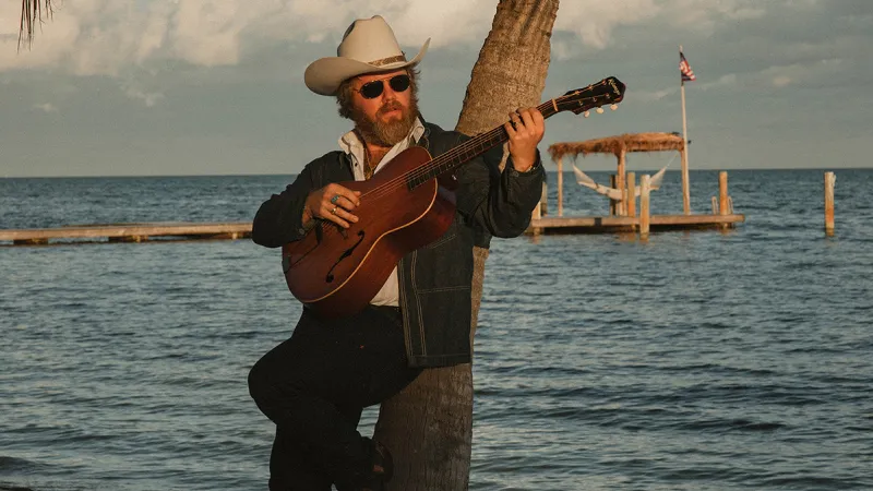 Country artist ERNEST plays guitar on the beach against a backdrop of a palm tree and late afternoon sky