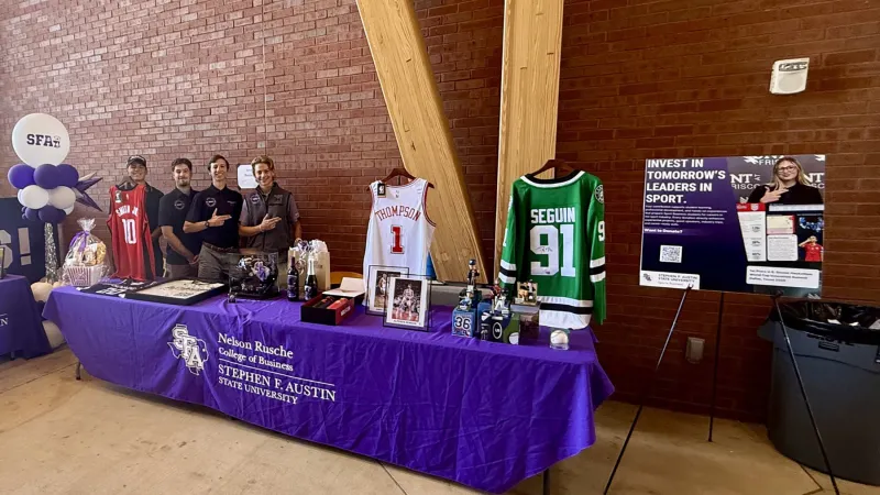 SFA Sports Business Society members stand behind a purple-clothed table displaying sports jerseys, photos, and other auction items.