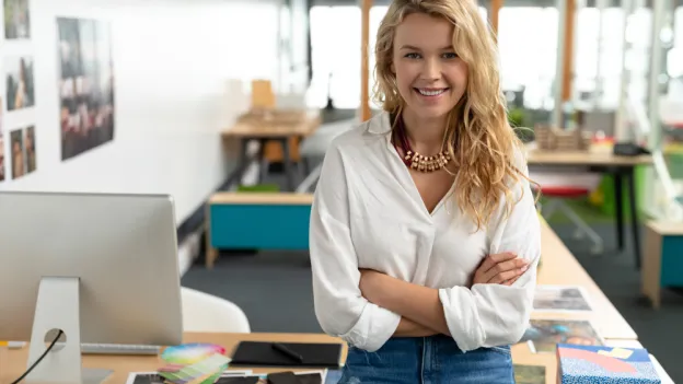 Portrait of Caucasian female graphic designer with arms crossed sitting at desk in a modern office. This is a casual creative start-up business office for a diverse team