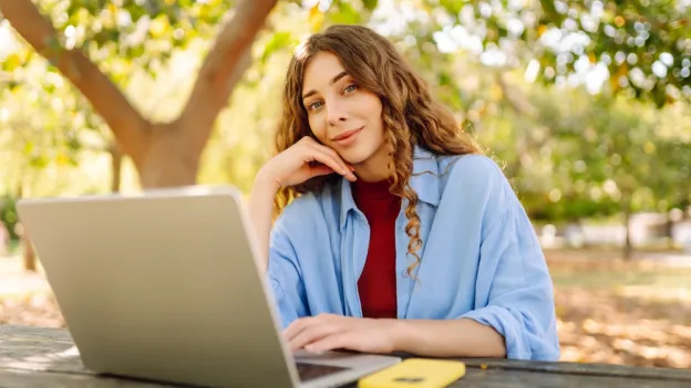 A young woman sits at a wooden table in a park using her laptop.