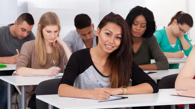 Portrait of smiling female college student sitting at desk with classmates in background