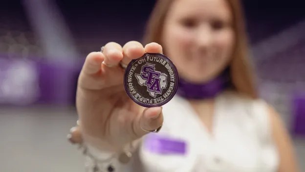 Hailey Crews displays the silver challenge coin she received. The coin displays the text, "Oh future bright 'neath the purple and white."