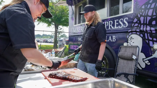 Axes and Embers team members prepare barbecue on a table next to the Lumberjack Express mobile food lab