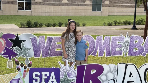 Arden and Ross Snyder are surrounded by glittery yard signs having the words "Welcome Back Jr Jacks"