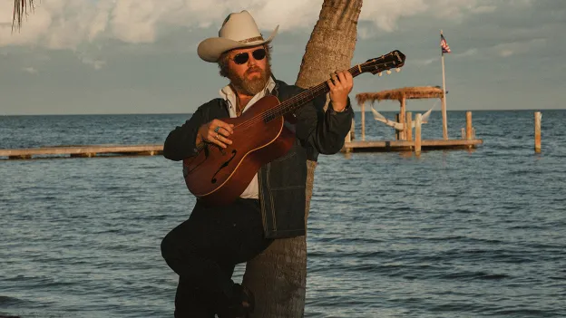 Country artist ERNEST plays guitar on the beach against a backdrop of a palm tree and late afternoon sky