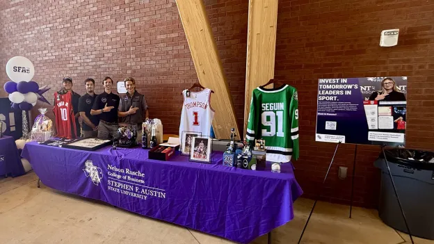 SFA Sports Business Society members stand behind a purple-clothed table displaying sports jerseys, photos, and other auction items.