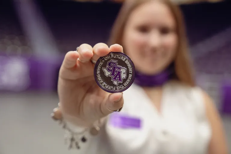 Hailey Crews displays the silver challenge coin she received. The coin displays the text, "Oh future bright 'neath the purple and white."