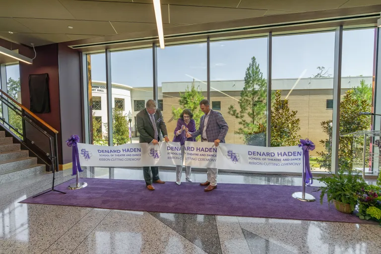 Standing on a purple carpet with a windowed backdrop of trees, Priscilla “Prissy” Abshier Sliva is assisted by Dr. Gary Wurtz and Dr. Neal Weaver in cutting a ceremonial ribbon.
