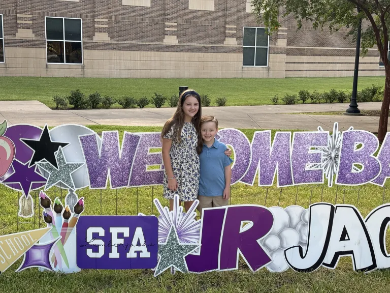 Arden and Ross Snyder are surrounded by glittery yard signs having the words "Welcome Back Jr Jacks"