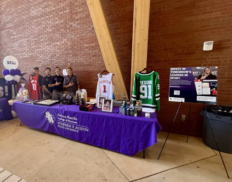 SFA Sports Business Society members stand behind a purple-clothed table displaying sports jerseys, photos, and other auction items.