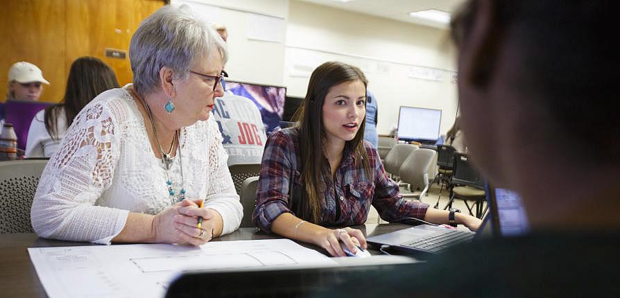 Several students and faculty member sit at a table and discuss a blueprint