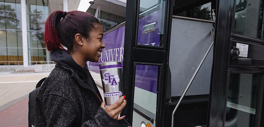 A student holding an SFA Lumberjacks tumbler smiles as she steps onto the shuttle bus