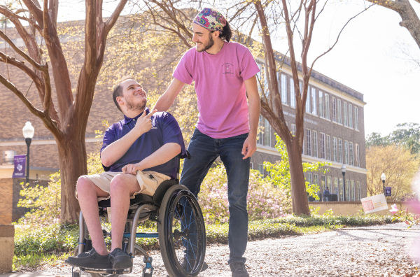 Two men outside. One standing and one sitting in a wheelchair.