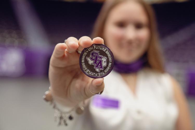 Hailey Crews displays the silver challenge coin she received. The coin displays the text, "Oh future bright 'neath the purple and white."