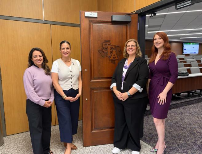 Four SFA alumnae are pictured standing in front of a wooden door stamped with the SFA logo