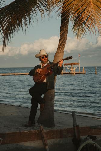 Country artist ERNEST plays guitar on the beach against a backdrop of a palm tree and late afternoon sky
