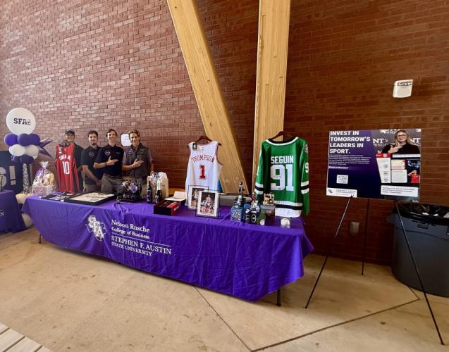 SFA Sports Business Society members stand behind a purple-clothed table displaying sports jerseys, photos, and other auction items.